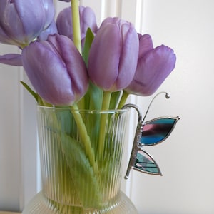 May include: A clear glass vase with a textured surface holds a bouquet of purple tulips. A stained glass butterfly with blue, pink and green accents is attached to the vase.