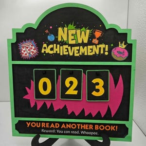 May include: A black and green wooden achievement tracker sign with the text NEW ACHIEVEMENT! and YOU READ ANOTHER BOOK! Reward: You can read. Whoopee. It features three flip-style number cards displaying 0, 2, and 3, with colorful icons.