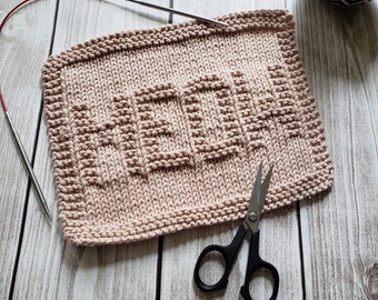 May include: A knitted dishcloth with the word "MOM" spelled out in the center. The dishcloth is a light pink color and is lying on a wooden surface. A pair of silver scissors with black handles are also on the surface.