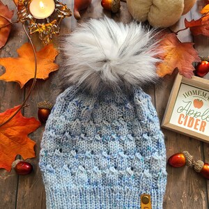 May include: A blue knitted hat with a white pom pom and a brown leather tag that says "Feltbun." The hat is surrounded by autumn leaves, acorns, and a white pumpkin.