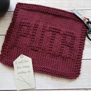 May include: A burgundy knitted dishcloth with a textured pattern. The dishcloth is lying on a wooden surface with a tag that reads "I could have been knitting something else".