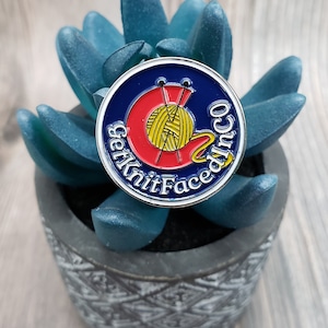 May include: A blue and red enamel pin with the text "Get Knit Faced" and a stylized yarn ball and knitting needles.