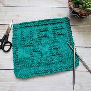 May include: A teal blue knitted dishcloth with a textured pattern. The dishcloth is lying on a wooden surface with two knitting needles and a pair of scissors.
