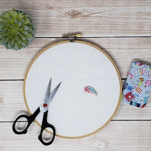 May include: A white embroidery hoop with a wooden frame, a pair of silver embroidery scissors, and a small tin box with a colorful pattern of sewing supplies.