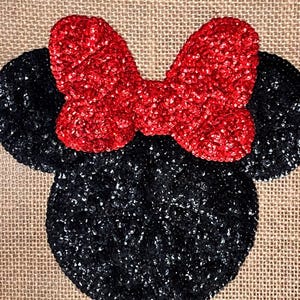 Op de afbeelding: Een decoratief jute stuk met een zwart, met pailletten bezet Mickey Mouse hoofd met twee ronde oren en een felrode, met pailletten bezette strik. De achtergrond is een natuurlijke bruine kleur.