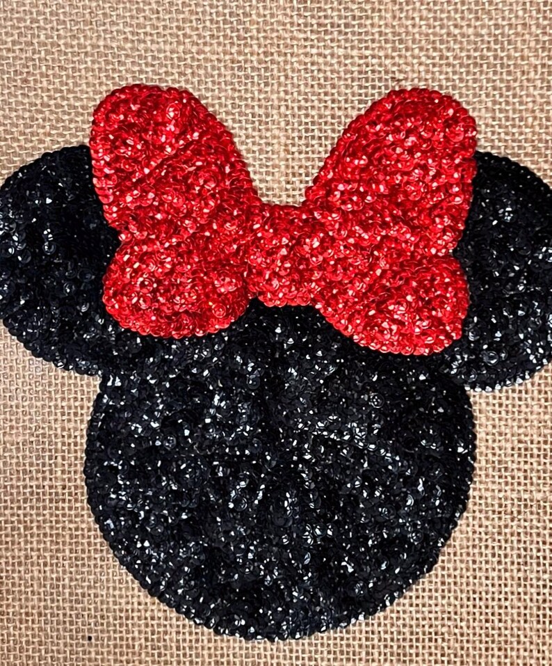 Op de afbeelding: Een decoratief jute stuk met een zwart, met pailletten bezet Mickey Mouse hoofd met twee ronde oren en een felrode, met pailletten bezette strik. De achtergrond is een natuurlijke bruine kleur.