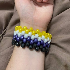 May include: A multi-coloured beaded bracelet with yellow, white, purple and black beads. The bracelet is worn on a person's wrist.