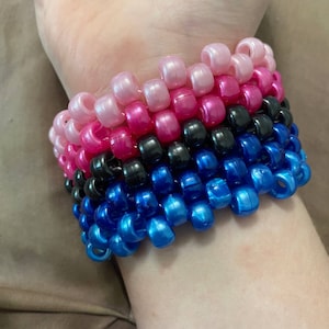 May include: A stack of colorful beaded bracelets in pink, hot pink, black, and blue. The bracelets are made of small, round beads that are strung together in a pattern.