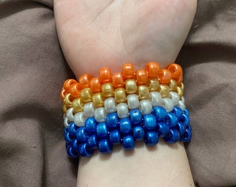 May include: A stack of colorful beaded bracelets in blue, white, gold, and orange. The bracelets are worn on a person's wrist.