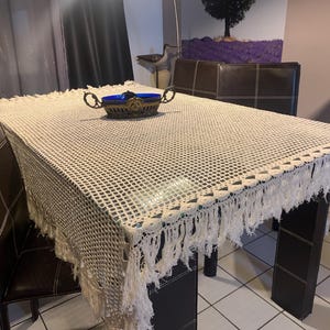 Peut inclure: Une nappe en crochet blanche avec des franges est posée sur une table en bois brun foncé avec quatre chaises. Un bol décoratif bleu et or est placé au centre de la table.