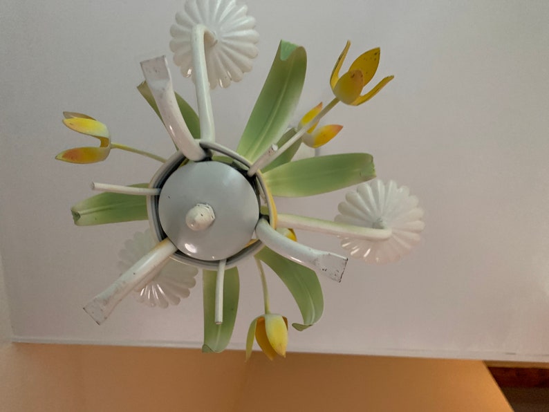 K&ouml;nnte beinhalten: Ein wei&szlig;er Metall-Kronleuchter mit gr&uuml;nen und gelben Blumenakzenten. Der Kronleuchter hat eine runde Basis und mehrere Arme, die sich nach au&szlig;en erstrecken, wobei jeder Arm mit einer Blume endet. Die Blumen sind eine Mischung aus Wei&szlig; und Gelb, einige haben einen gr&uuml;nen Stiel.