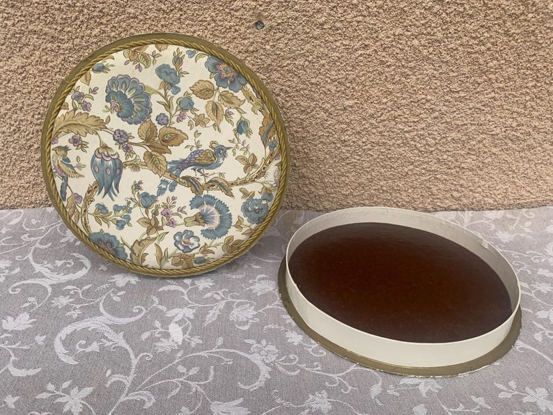 May include: A round, floral patterned box with a gold trim. The lid is open and the inside is lined with a brown fabric.
