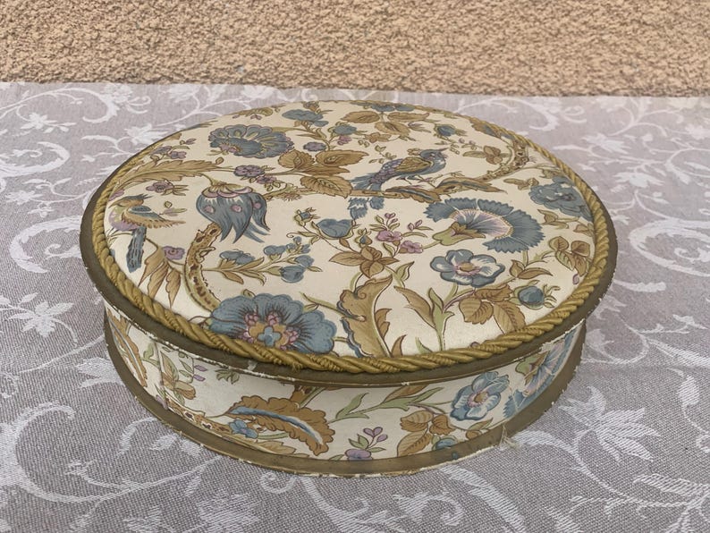 May include: A round, vintage trinket box with a floral pattern in shades of blue, brown, and pink. The box has a gold trim and a rope-like design around the edge.