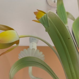 K&ouml;nnte beinhalten: Nahaufnahme eines Metall-Kronleuchters mit gelben und gr&uuml;nen Blumenakzenten. Der Kronleuchter ist aus wei&szlig;em Metall gefertigt und hat ein florales Design.