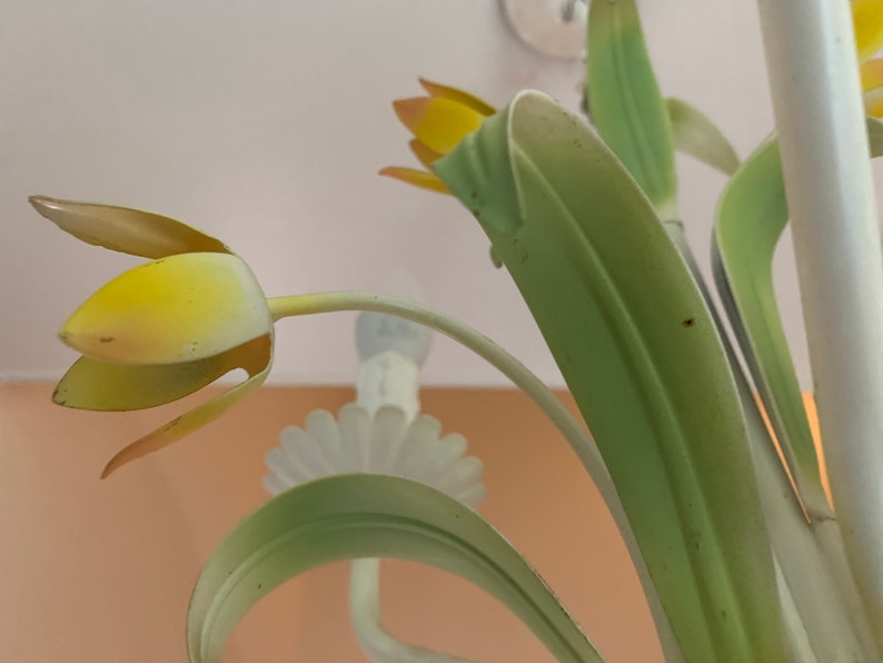 K&ouml;nnte beinhalten: Nahaufnahme eines Metall-Kronleuchters mit gelben und gr&uuml;nen Blumenakzenten. Der Kronleuchter ist aus wei&szlig;em Metall gefertigt und hat ein florales Design.