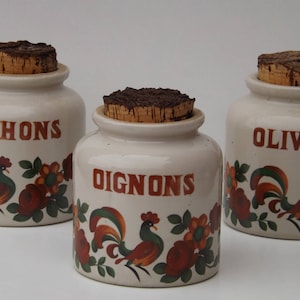 Peut inclure: Trois pots en céramique avec des couvercles en liège et des illustrations de coqs. Les pots sont étiquetés "Cornichons", "Oignons" et "Olives".