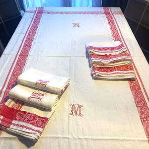 May include: A white linen tablecloth with red embroidered borders and initials. The tablecloth is folded and stacked on a table with two chairs.
