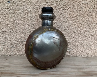 Könnte beinhalten: Eine vintage Metallflasche mit runder Form und einem dunkelbraunen Verschluss. Die Flasche hat ein abgenutztes, gealtertes Aussehen mit einer metallischen Oberfläche und einem Messingband um die Mitte.