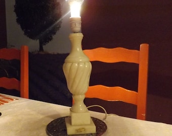 Peut inclure: Une lampe de table en marbre blanc avec un motif en spirale et une ampoule transparente. La lampe est posée sur une table avec une nappe blanche et un fond brun foncé.