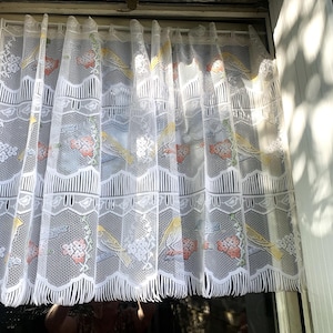 以下が含まれることがあります： 黄色い鳥と赤い花の繰り返し模様が施された白いレースの窓カーテン。 カーテンはスカラップエッジで、窓に吊るされています。