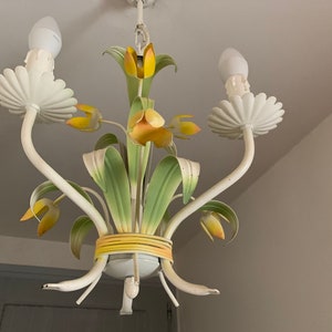 K&ouml;nnte beinhalten: Ein wei&szlig;er Metall-Kronleuchter mit gelben und gr&uuml;nen floralen Akzenten. Der Kronleuchter hat drei Lichter und eine dekorative Basis.