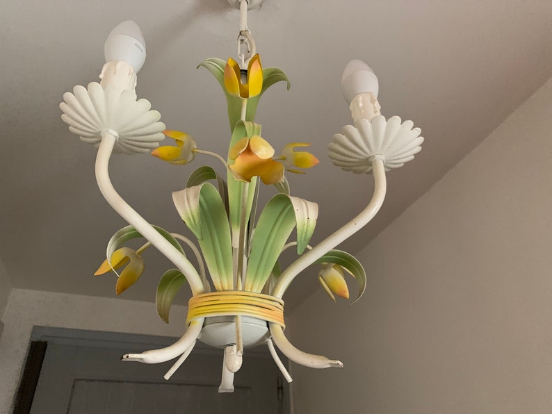 K&ouml;nnte beinhalten: Ein wei&szlig;er Metall-Kronleuchter mit gelben und gr&uuml;nen floralen Akzenten. Der Kronleuchter hat drei Lichter und eine dekorative Basis.