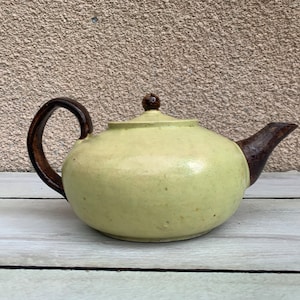 以下が含まれることがあります： 茶色の取っ手と注ぎ口が付いた、薄い緑色のセラミック製のティーポット。ティーポットは斑点のある釉薬と丸みを帯びたボディをしています。
