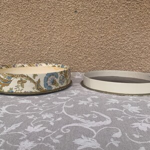 May include: A round, floral patterned box with a white background and blue, brown, and green accents. The box is open and the lid is sitting next to it.