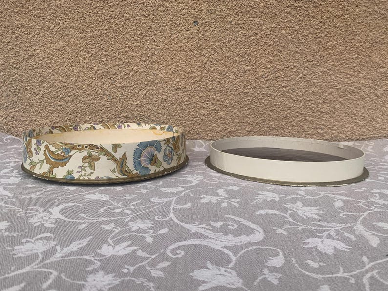May include: A round, floral patterned box with a white background and blue, brown, and green accents. The box is open and the lid is sitting next to it.