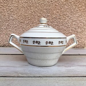 以下が含まれることがあります： 金色の縁取りと花柄の白い陶器製の砂糖入れ。蓋の上にはノブがあります。