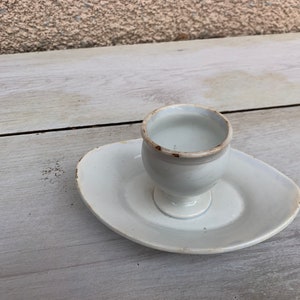 以下が含まれることがあります： 白いセラミック製の卵立てと小さな受け皿。卵立ては丸みを帯びた形をしていて、小さな足がついています。受け皿は楕円形です。