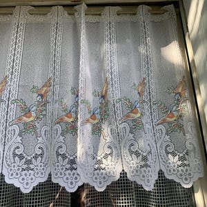 以下が含まれることがあります： 枝にとまっている鳥の繰り返し模様が特徴の白いレースのカーテン。鳥は茶色、青、黄色の色をしています。カーテンの下端はスカラップのデザインになっています。