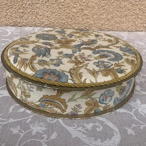 May include: A round, vintage storage box with a floral pattern in shades of blue, brown, and white. The box has a gold trim around the top edge.