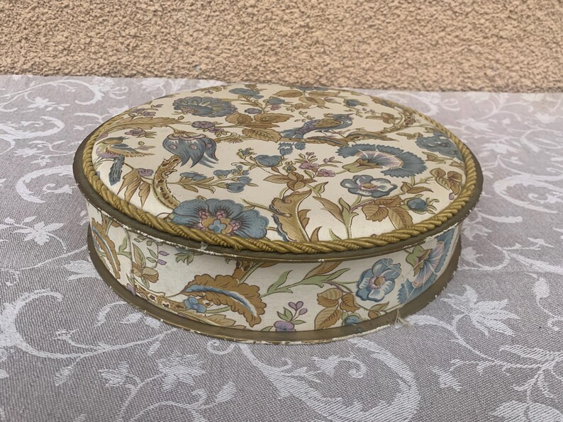 May include: A round, vintage storage box with a floral pattern in shades of blue, brown, and white. The box has a gold trim around the top edge.