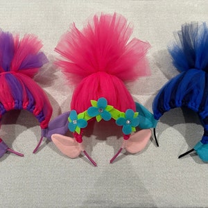 May include: Three colorful troll hair headbands with tulle hair and felt ears. The headbands are pink, purple, and blue.