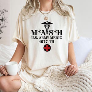 Może przedstawiać: Kremowa koszulka z czarnym napisem "M*A*S*H U.S. ARMY MEDIC 4077 TH", symbolem medycznym i czerwonym krzyżem. Osoba ma na sobie koszulkę i trzyma biały kubek.
