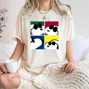 Peut inclure: T-shirt crème avec un motif de Snoopy portant des lunettes de soleil dans quatre carrés de couleurs différentes : vert, rouge, bleu et jaune. Le t-shirt est de style décontracté et confortable.
