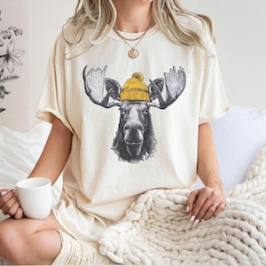 Könnte beinhalten: Cremefarbenes T-Shirt mit einer detaillierten Illustration eines Elchs mit gelber Mütze. Der Elch hat ein großes Geweih und einen ernsten Gesichtsausdruck. Das Shirt hat eine lockere Passform, geeignet für Freizeitkleidung.