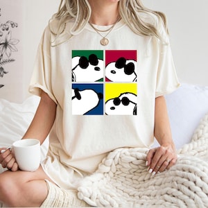 Peut inclure: T-shirt crème avec un motif de Snoopy portant des lunettes de soleil dans quatre carrés de couleurs différentes : vert, rouge, bleu et jaune. Le t-shirt est de coupe ample et décontractée.