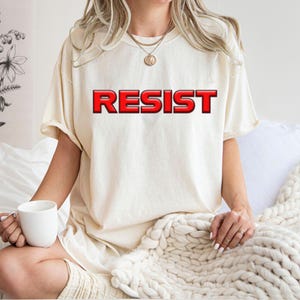 Puede incluir: Camiseta color crema con la palabra "RESIST" en letras rojas en relieve. La persona lleva la camiseta y está sentada en una cama con una manta blanca, sosteniendo una taza blanca. En el fondo se ve un estampado floral.