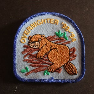May include: Blue and yellow embroidered patch with a sleeping beaver on a branch, surrounded by green leaves. The text "OVERNIGHTER 93-94" is stitched in yellow around the edge.