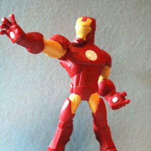 May include: A red and yellow Iron Man action figure stands on a gray base. The figure is posed with one arm outstretched and the other hand clenched into a fist.