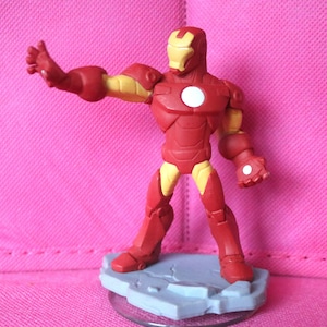 May include: A red and yellow Iron Man figurine. The figure is posed on a grey base with an arm outstretched. The figure has a white circle on its chest and a yellow face mask.