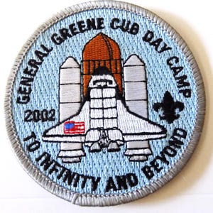 May include: Embroidered patch with a light blue background and a silver border. The patch features a space shuttle design with the American flag and the text "GENERAL GREENE CUB DAY CAMP 2002 TO INFINITY AND BEYOND". A black fleur-de-lis is also present.
