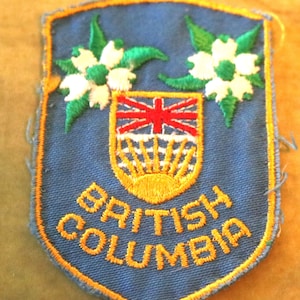 May include: A blue embroidered patch with a yellow border. The patch features a crest with a red cross on a white background, a yellow sun, and two white flowers with green leaves. The text "BRITISH COLUMBIA" is embroidered in yellow below the crest.