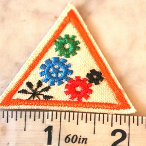 May include: A triangular Girl Scout patch with an orange border and five colorful embroidered gears. The patch is about 1 1/2 inches wide.