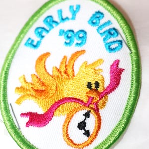 May include: A green oval embroidered patch with a yellow bird holding a pink ribbon and a clock. The text "Early Bird '99" is written in blue around the edge of the patch.