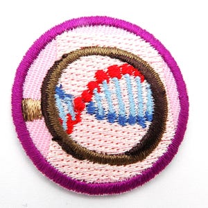 May include: Round embroidered patch featuring a DNA double helix design. The patch has a light pink background with a brown outline around the DNA. The DNA is depicted in red and blue. The patch is bordered by a dark pink and purple trim.