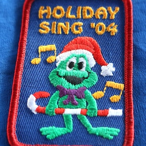 May include: A blue embroidered patch with red stitching featuring a green frog wearing a red and white Santa hat, holding a candy cane, and surrounded by musical notes. The text "HOLIDAY SING '04" is embroidered in yellow.
