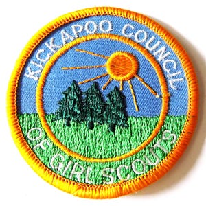 May include: Embroidered patch with the text "Kickapoo Council" and "Girl Scouts" in a circular design. The patch features a blue sky, green grass, three green trees, and an orange sun. The border is orange.